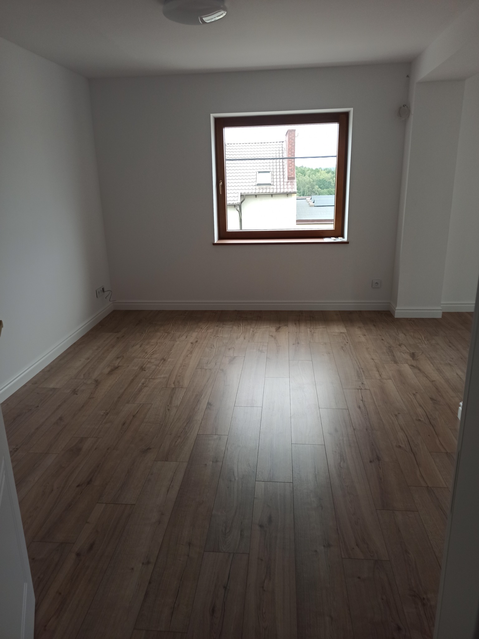 Pusty pokój z nową podłogą z paneli w odcieniach brązu, widok na okno z białymi ścianami i listwami przypodłogowymi. W tle widoczny budynek z czerwonym kominem.