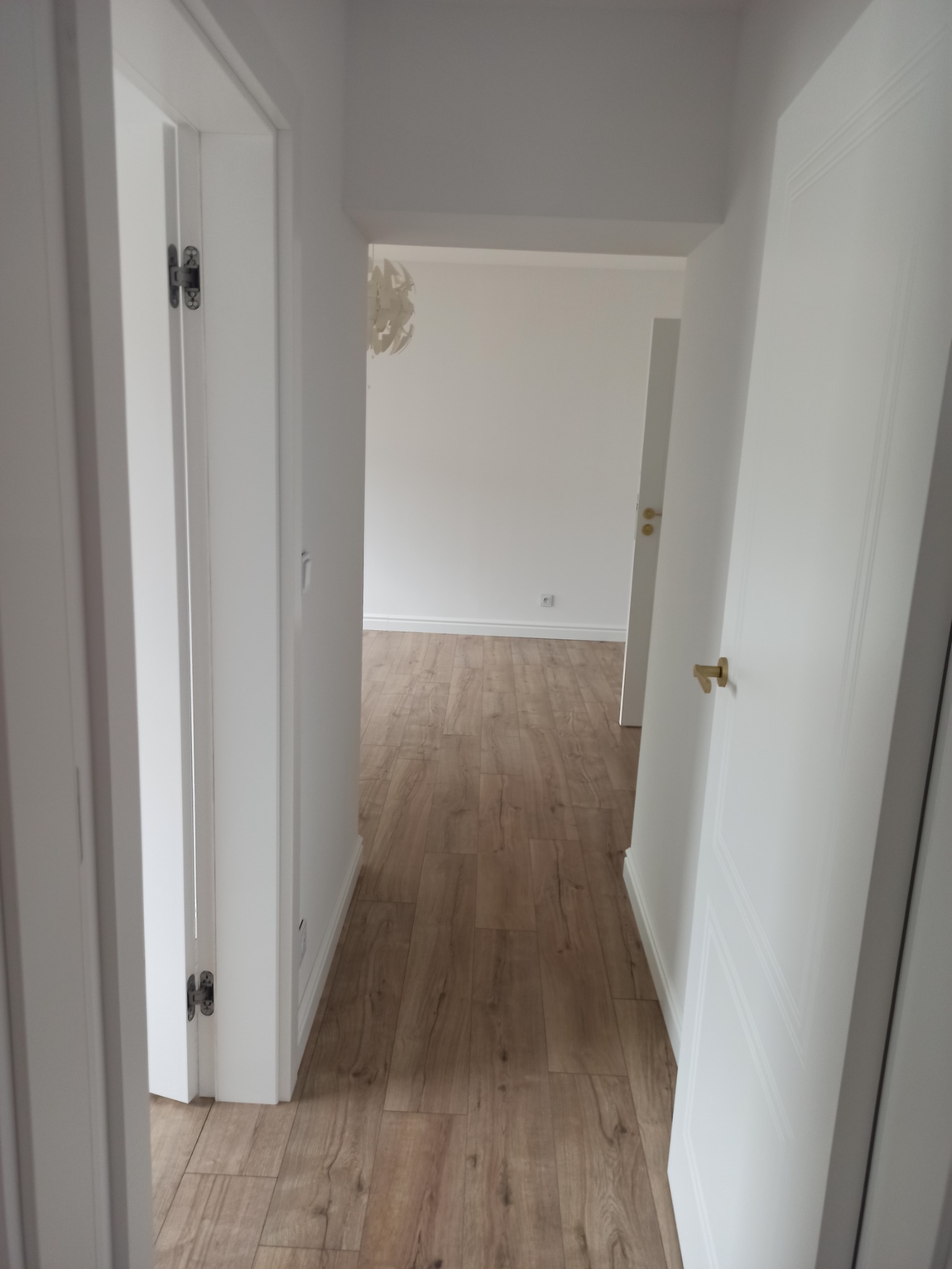 Widok z korytarza na odnowione wnętrze z drewnianą podłogą i białymi drzwiami z mosiężnymi klamkami, w tle widoczny dekoracyjny żyrandol.