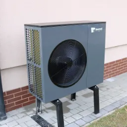 Szara jednostka zewnętrzna z widocznym logo Kensol HVAC, zamontowana na czarnych wspornikach przy ścianie budynku z ceglaną podmur&oacute;wką i szarą kostką brukową.