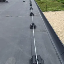 Widok z góry na płaski dach z zamontowanym systemem odgromowym: przewód biegnie wzdłuż dachu, podtrzymywany przez czarne plastikowe uchwyty w kształcie ściętego ostrosłupa, w tle widoczna zieleń...