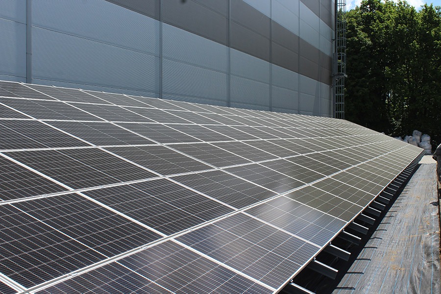 Rozległa instalacja paneli słonecznych na dachu przemysłowym, widoczne elementy konstrukcyjne i fragment budynku z drabinką po lewej stronie, drzewa w tle.