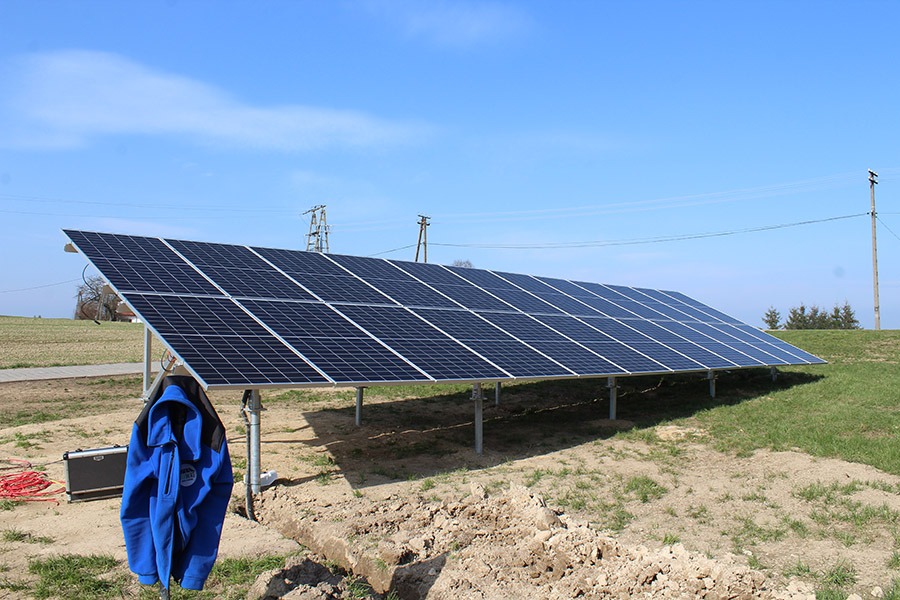 Panel fotowoltaiczny na metalowych wspornikach, na trawiastym terenie z widoczną skrzynką narzędziową i powieszoną niebieską kurtką roboczą, na tle błękitnego nieba.