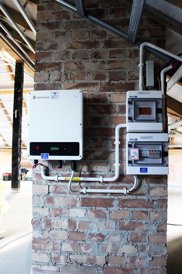 Biały falownik firmy Goodwe zamontowany na ceglanej ścianie w pomieszczeniu w Tarnowie, z widocznymi przewodami i rozdzielnicą elektryczną, w tle konstrukcja dachu.