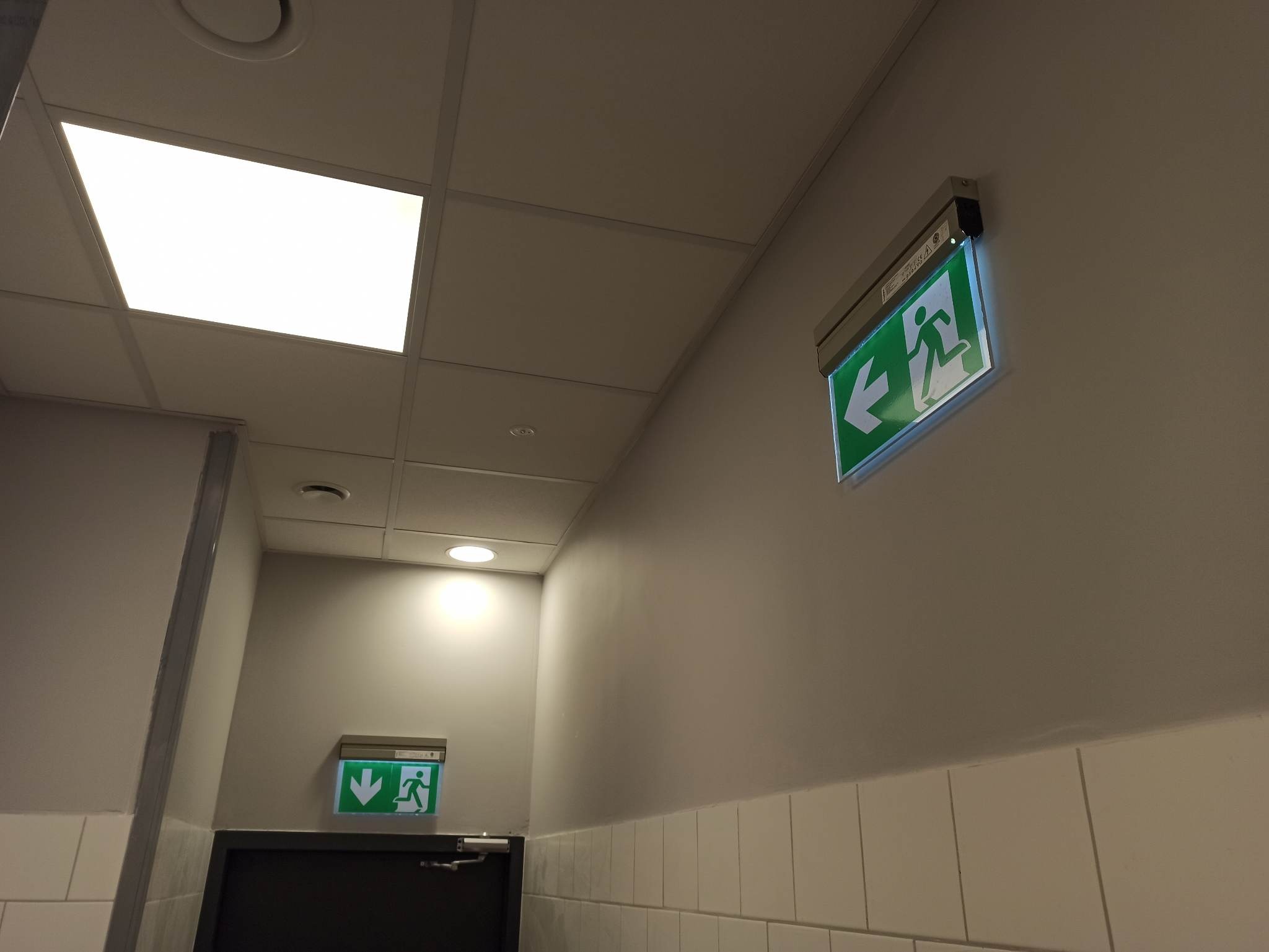 Korytarz z dwoma podświetlanymi znakami ewakuacyjnymi: jeden wskazuje kierunek w lewo, drugi w dół nad drzwiami.