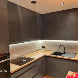 Nowoczesna kuchnia z szarymi matowymi szafkami, drewnianym blatem, czarnym zlewem i płytą indukcyjną, oświetleniem LED podszafkowym i szarą glazurą na ścianie.