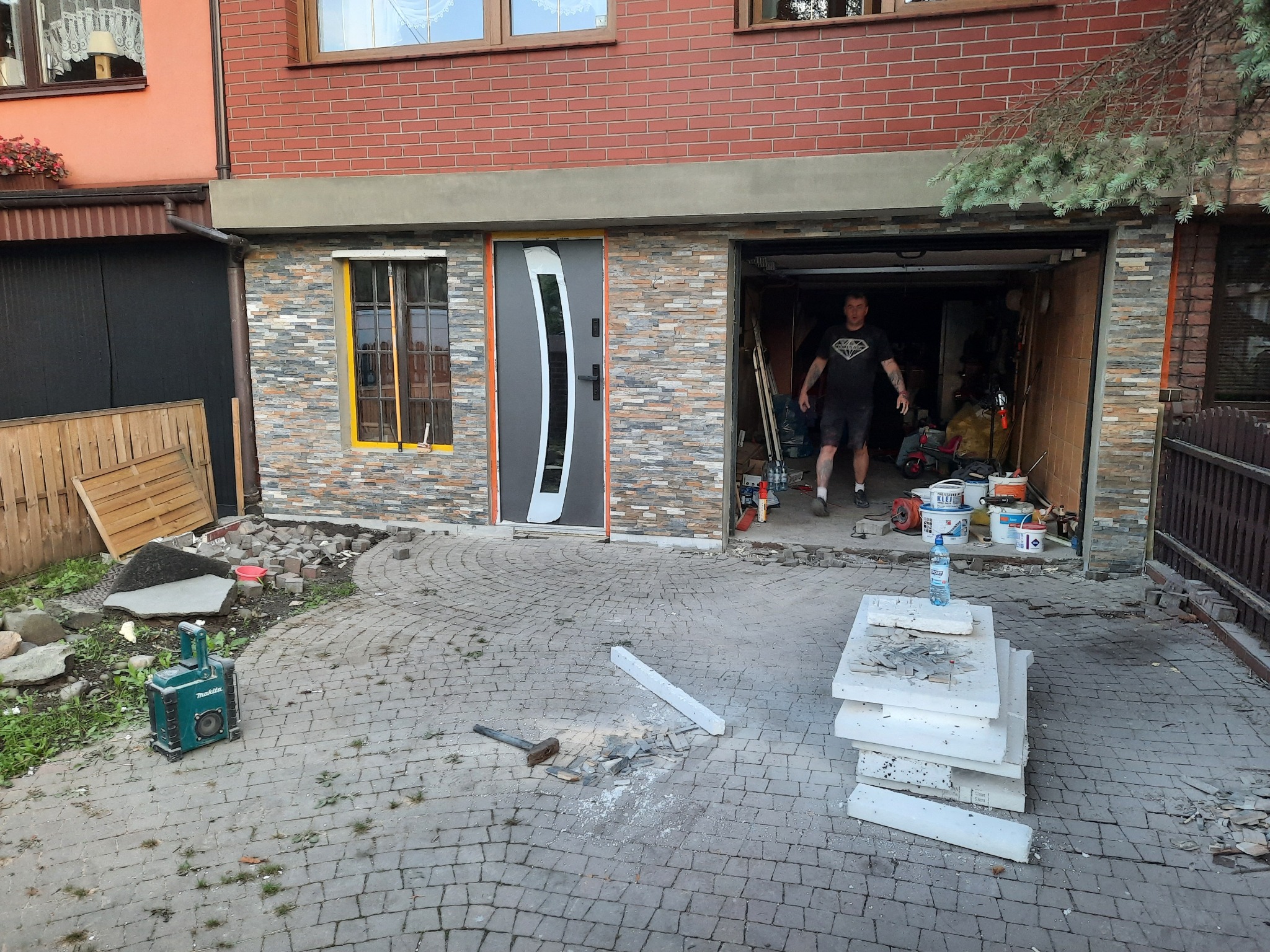 Układanie kamienia elewacyjnego na zewnętrznej ścianie domu, widoczne narzędzia, resztki materiału, oraz pracownik w tle w otwartym garażu.