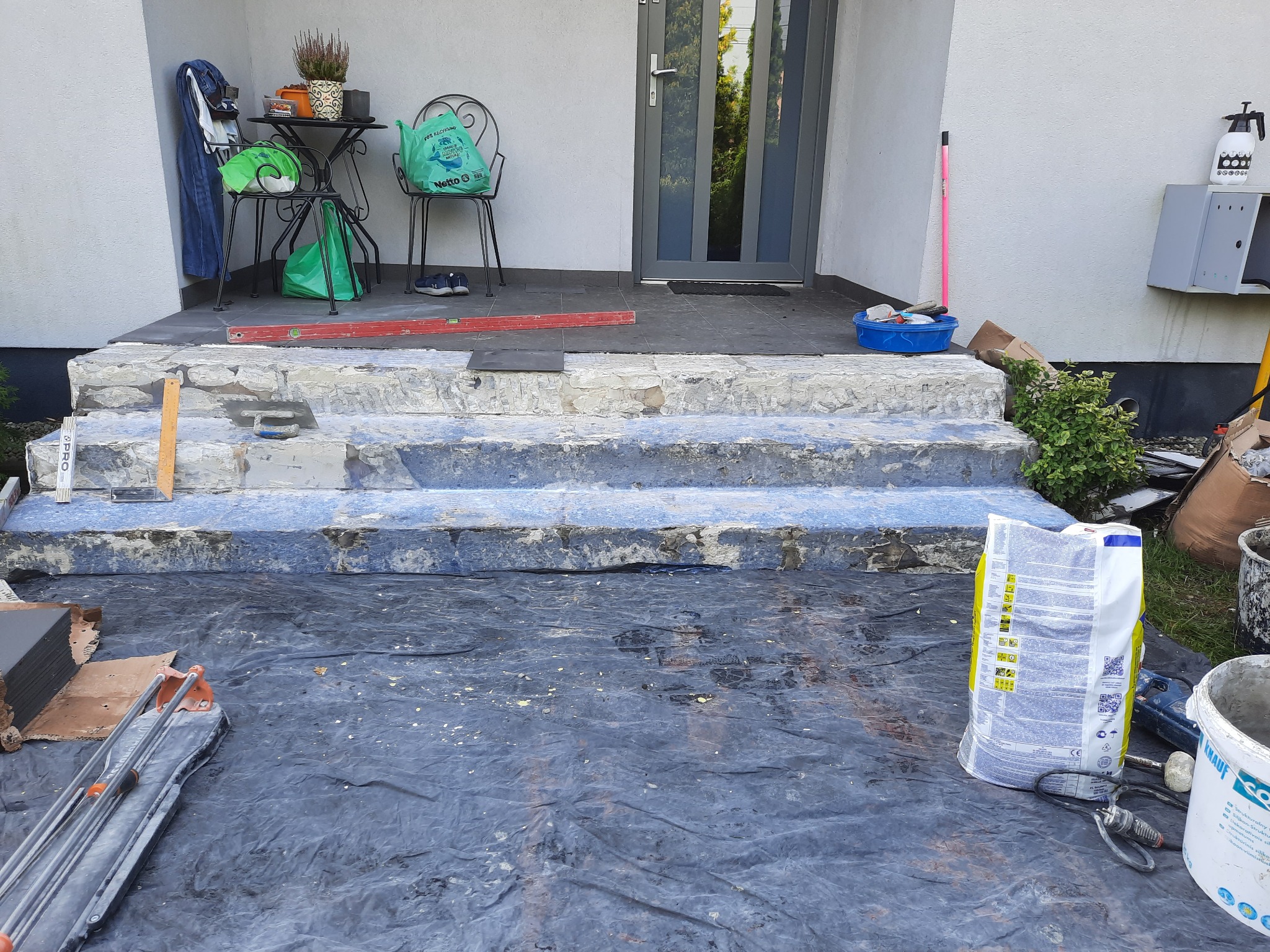 Przygotowanie do układania płytek na schodach wejściowych: widoczne narzędzia glazurnicze, poziomica, paczka kleju Knauf oraz resztki starej posadzki.