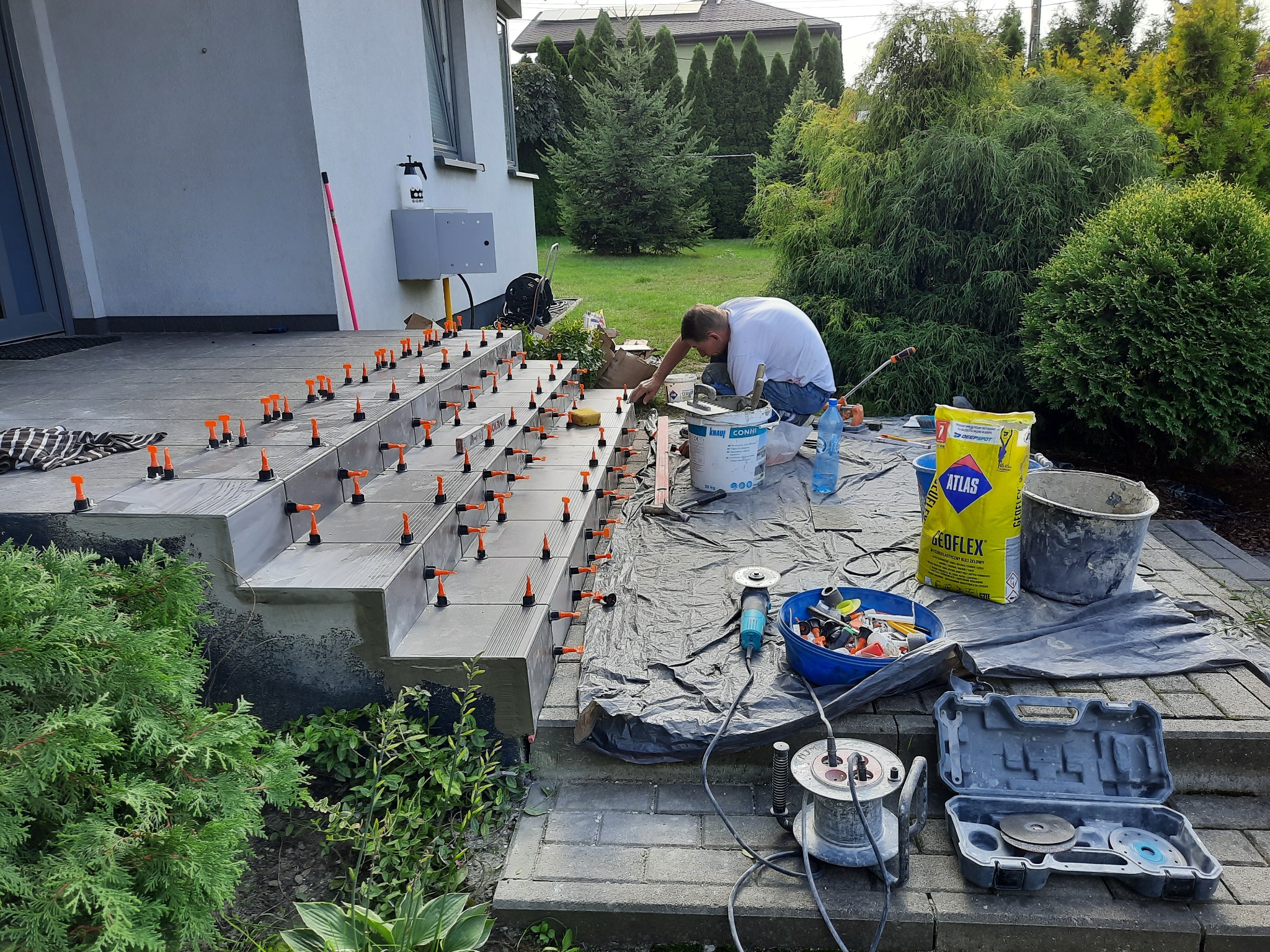 Układanie płytek na schodach zewnętrznych, widoczne poziomice i system poziomowania płytek, w tle dom i zieleń.