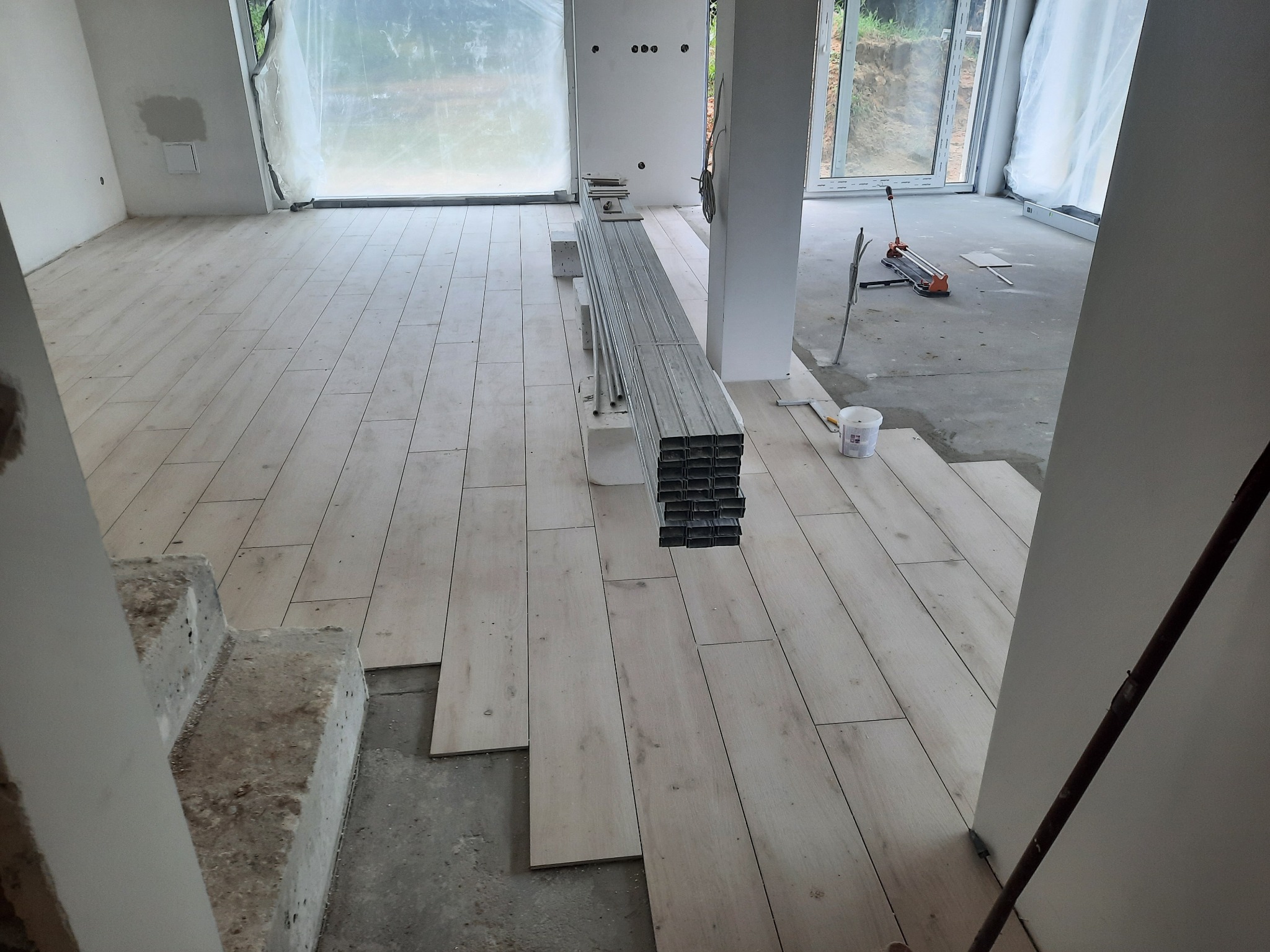 Wnętrze w trakcie układania podłogi z paneli imitujących drewno; widoczne niedokończone schody, stos profili metalowych na podkładkach, narzędzia do cięcia płytek w tle.