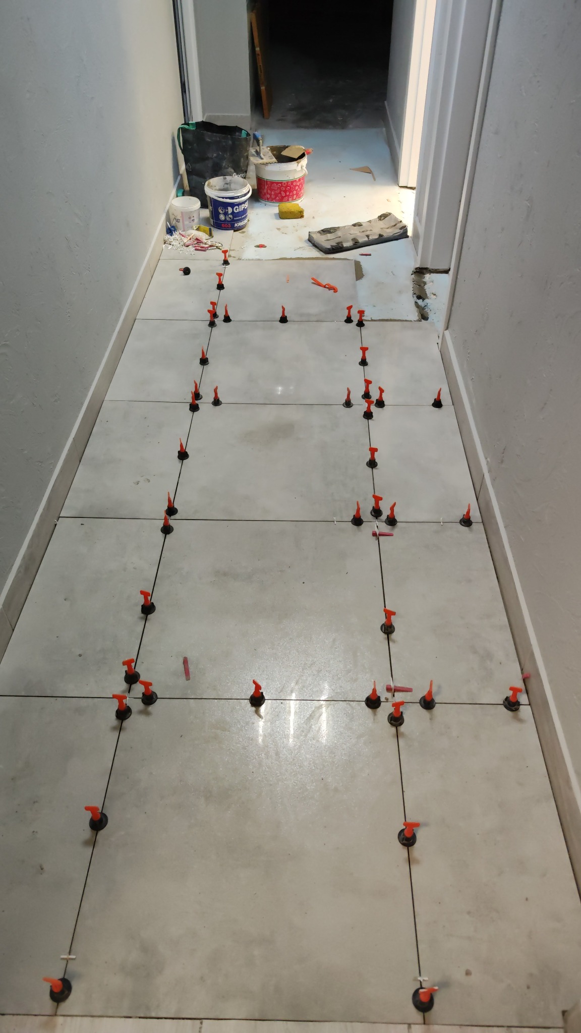 Kładzenie jasnych, kwadratowych płytek podłogowych w wąskim korytarzu, z użyciem systemu poziomowania z czarnymi podstawami i pomarańczowymi klinami, widoczne wiaderka z zaprawą i narzędzia...