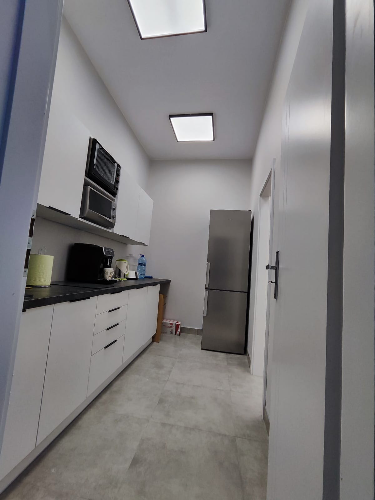 Nowoczesna kuchnia w odcieniach szarości i bieli z czarnym blatem, oświetlona dwoma kwadratowymi lampami sufitowymi, z widoczną zabudową kuchenną, sprzętem AGD i szarą podłogą.