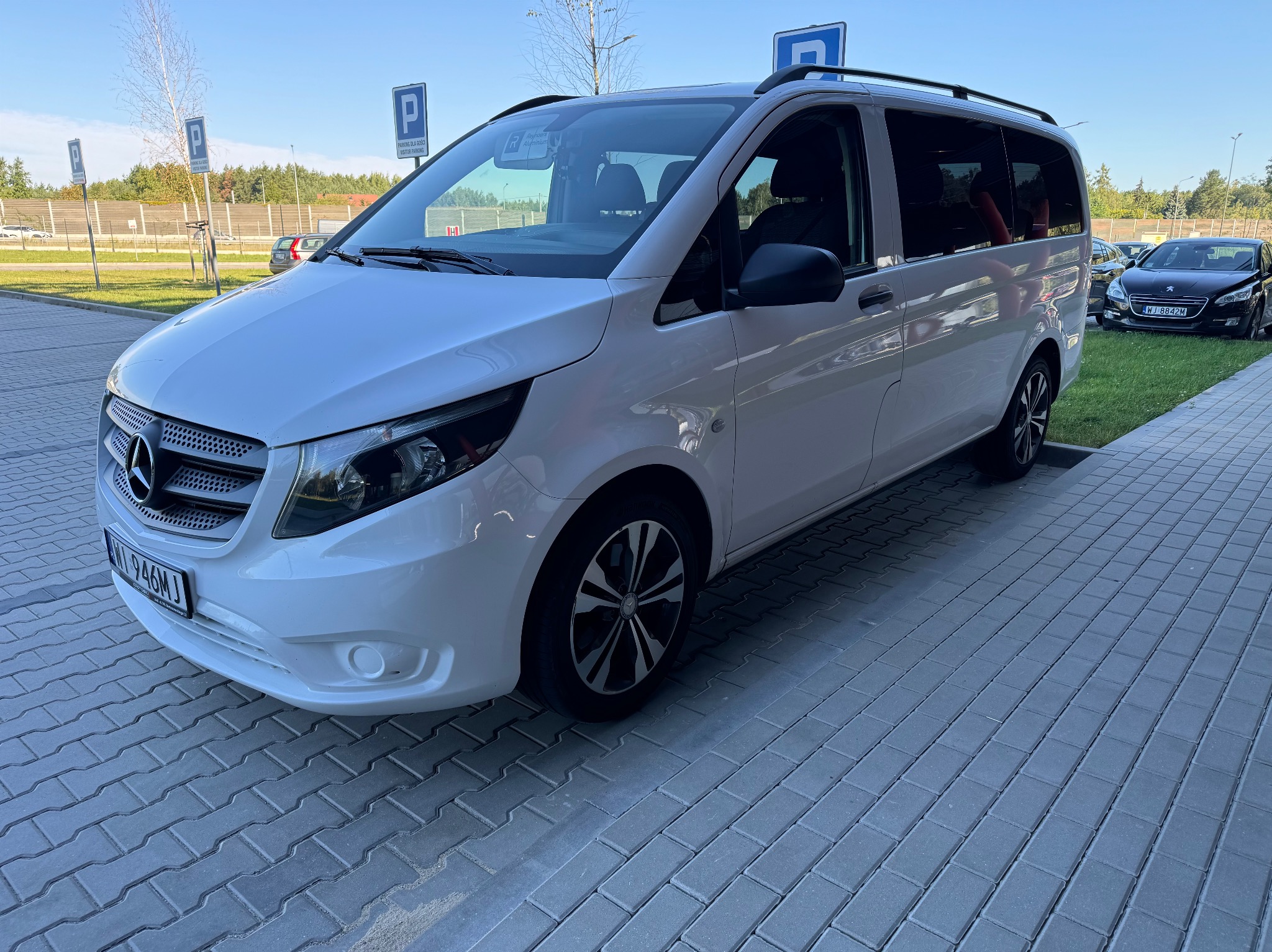 Biały Mercedes Vito z bagażnikiem dachowym zaparkowany na parkingu, widoczne tablice rejestracyjne i oznaczenia parkingowe.