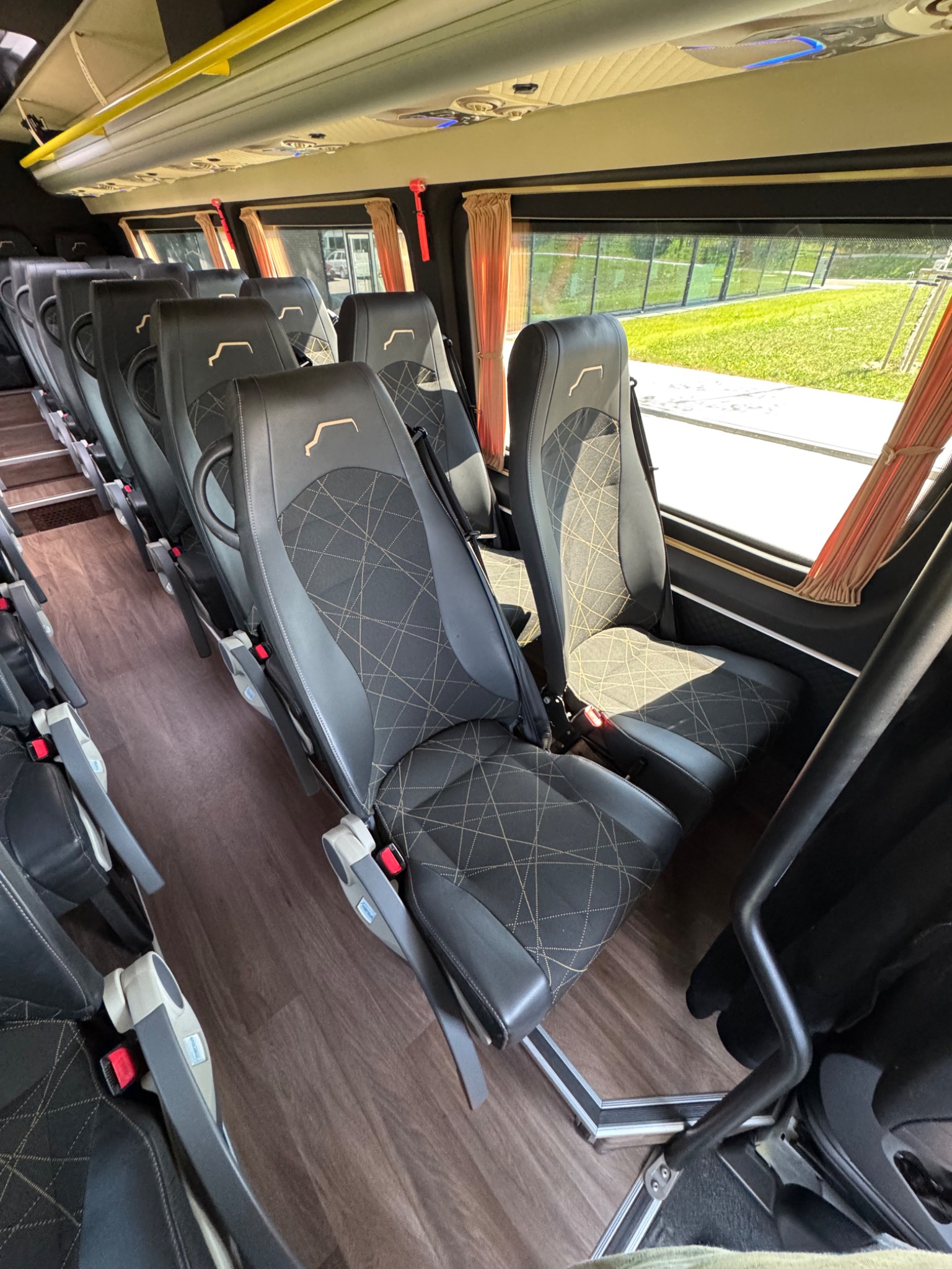 Wnętrze autobusu z rzędami czarnych skórzanych foteli z geometrycznym wzorem, widok z boku, jasne światło dzienne wpadające przez okna z pomarańczowymi zasłonami.