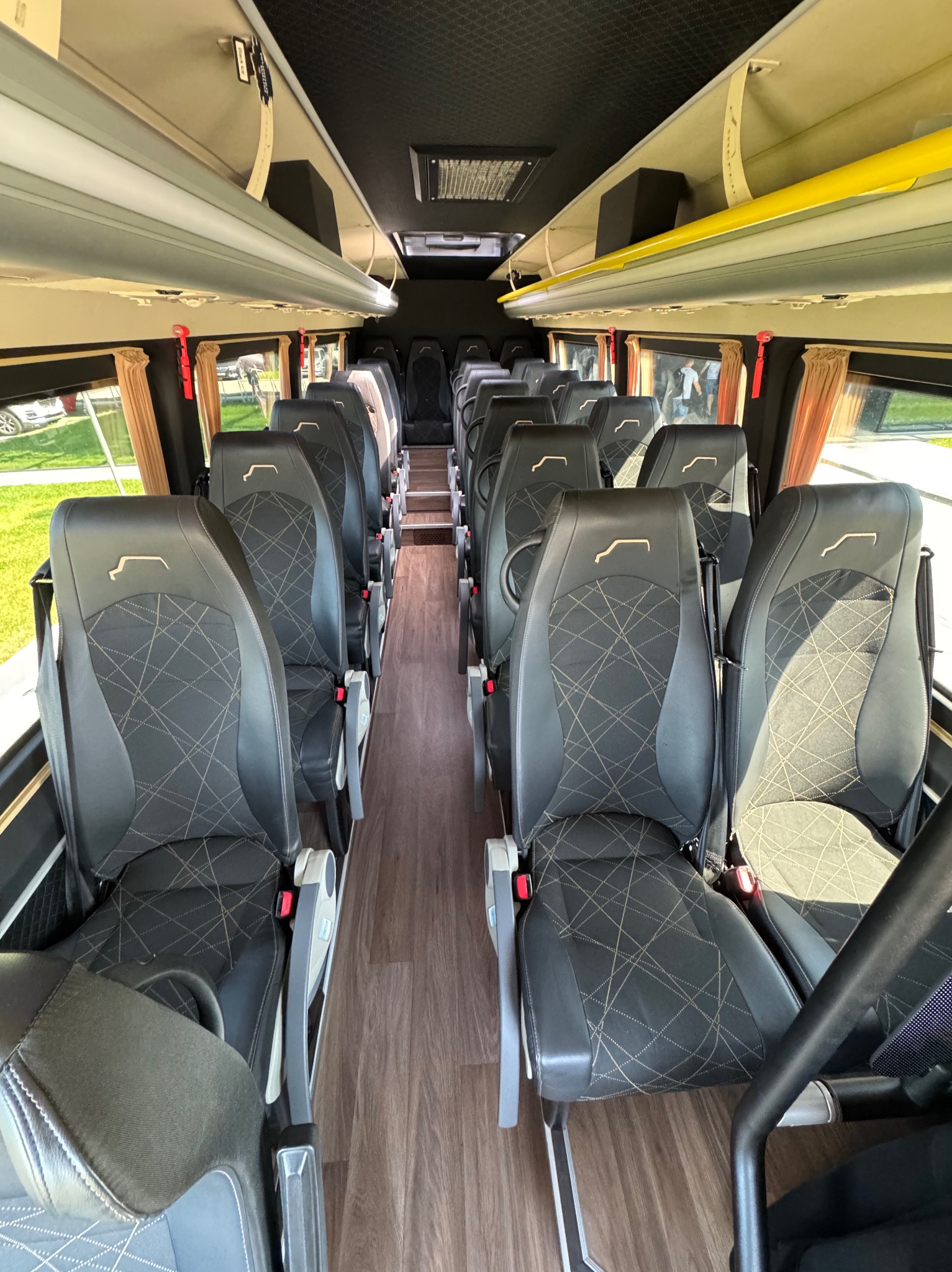 Wnętrze autobusu z rzędami czarnych skórzanych siedzeń z geometrycznym wzorem, drewnopodobna podłoga i beżowe zasłony w oknach.