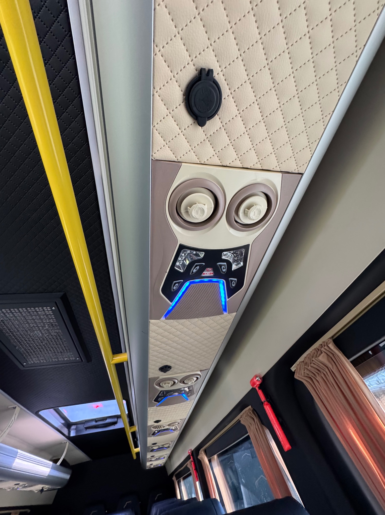 Wnętrze autobusu turystycznego: beżowa pikowana podsufitka z gniazdem USB, panel sterowania klimatyzacją z niebieskim podświetleniem, żółty uchwyt, brązowe zasłony w oknach.