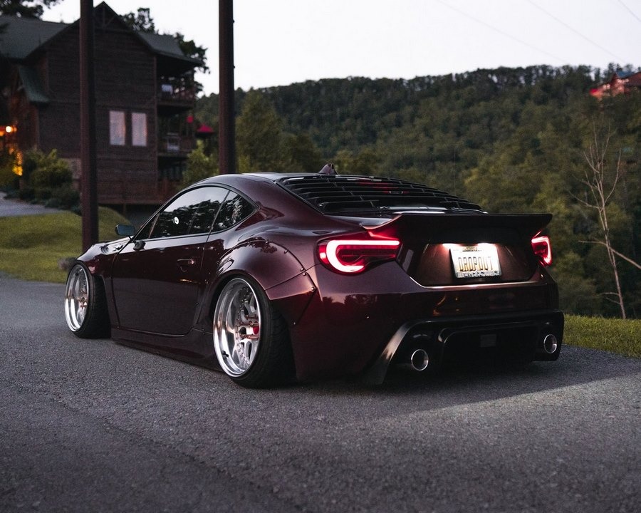 Burgundowy, zmodyfikowany samochód sportowy z szerokim body kitem i chromowanymi felgami, zaparkowany na drodze z widokiem na dom w stylu góralskim i zalesione wzgórze.