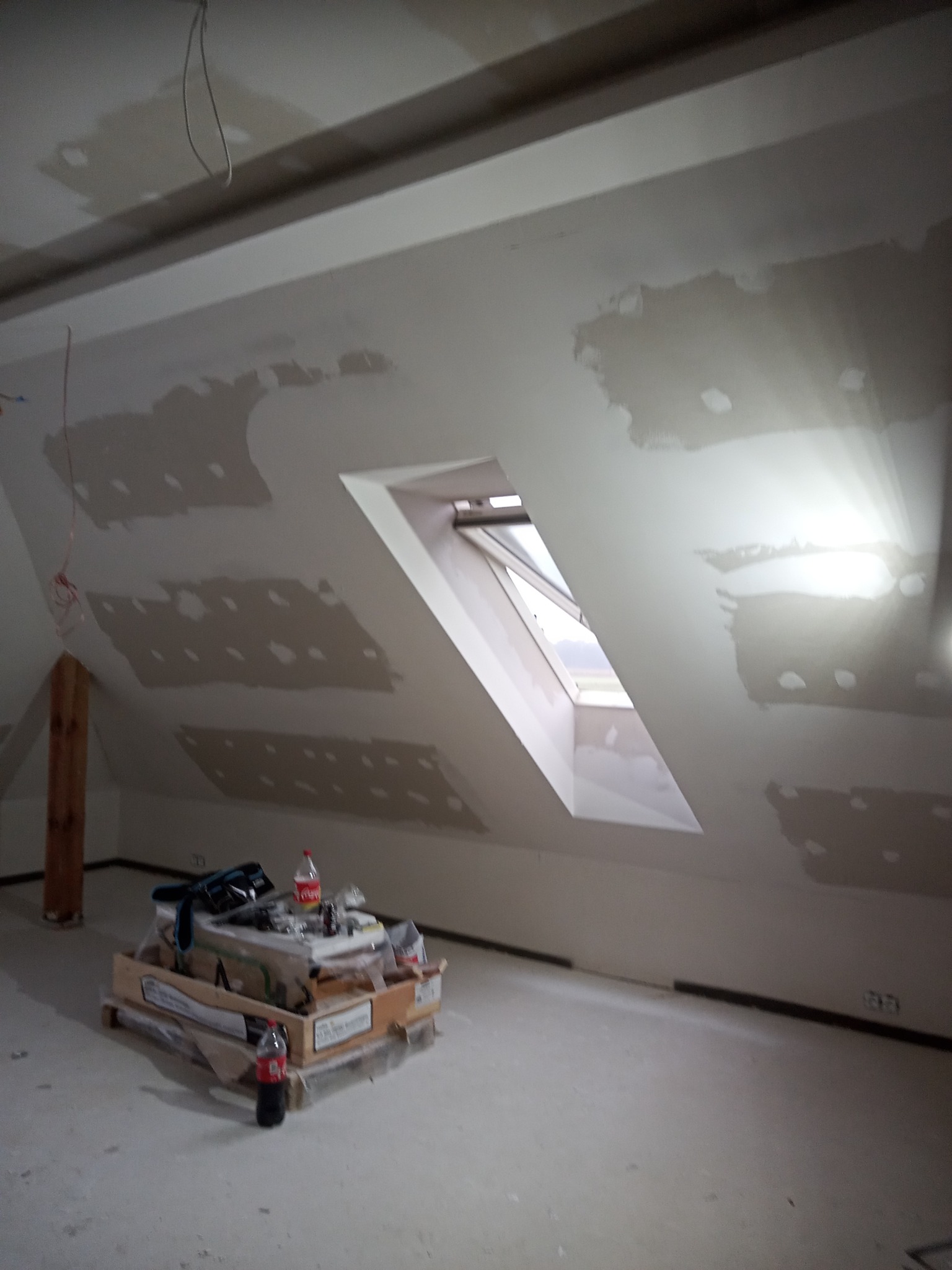 Poddasze w trakcie prac wykończeniowych z oknem dachowym, widoczne szpachlowane miejsca na ścianach i stropie, paleta z narzędziami i materiałami budowlanymi w centralnej części pomieszczenia.
