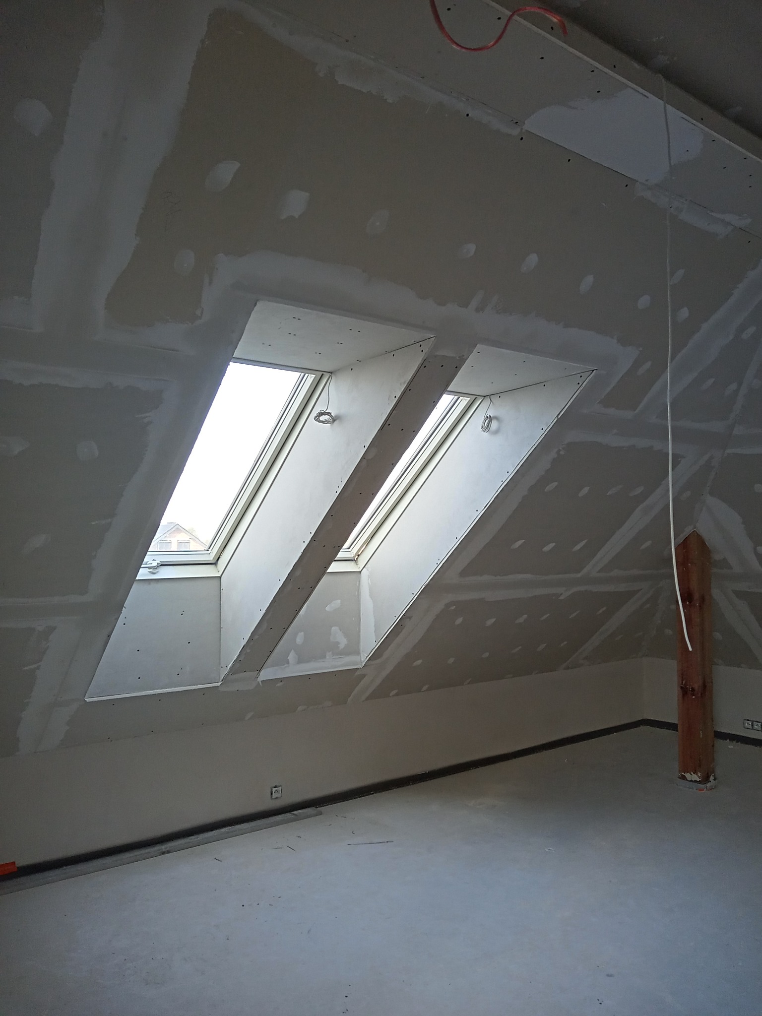 Poddasze w trakcie prac wykończeniowych: zamontowane okna dachowe, ściany i sufit pokryte płytami, widoczne łączenia płyt przed szpachlowaniem, drewniany słup nośny.