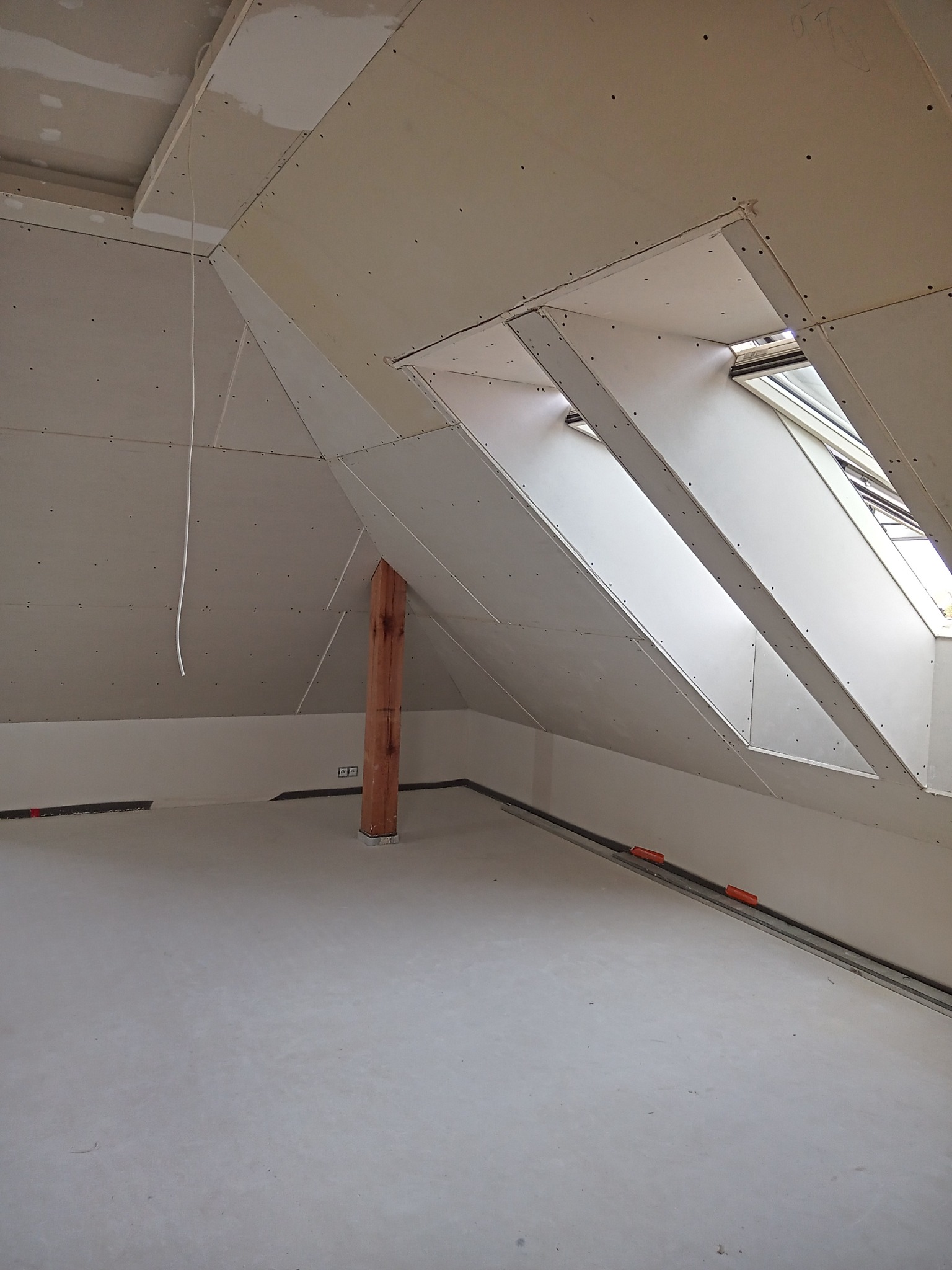 Poddasze w trakcie prac wykończeniowych, widoczne zamontowane płyty gipsowo-kartonowe na skosach dachu z oknami, drewniany słup nośny i świeżo wylana podłoga.