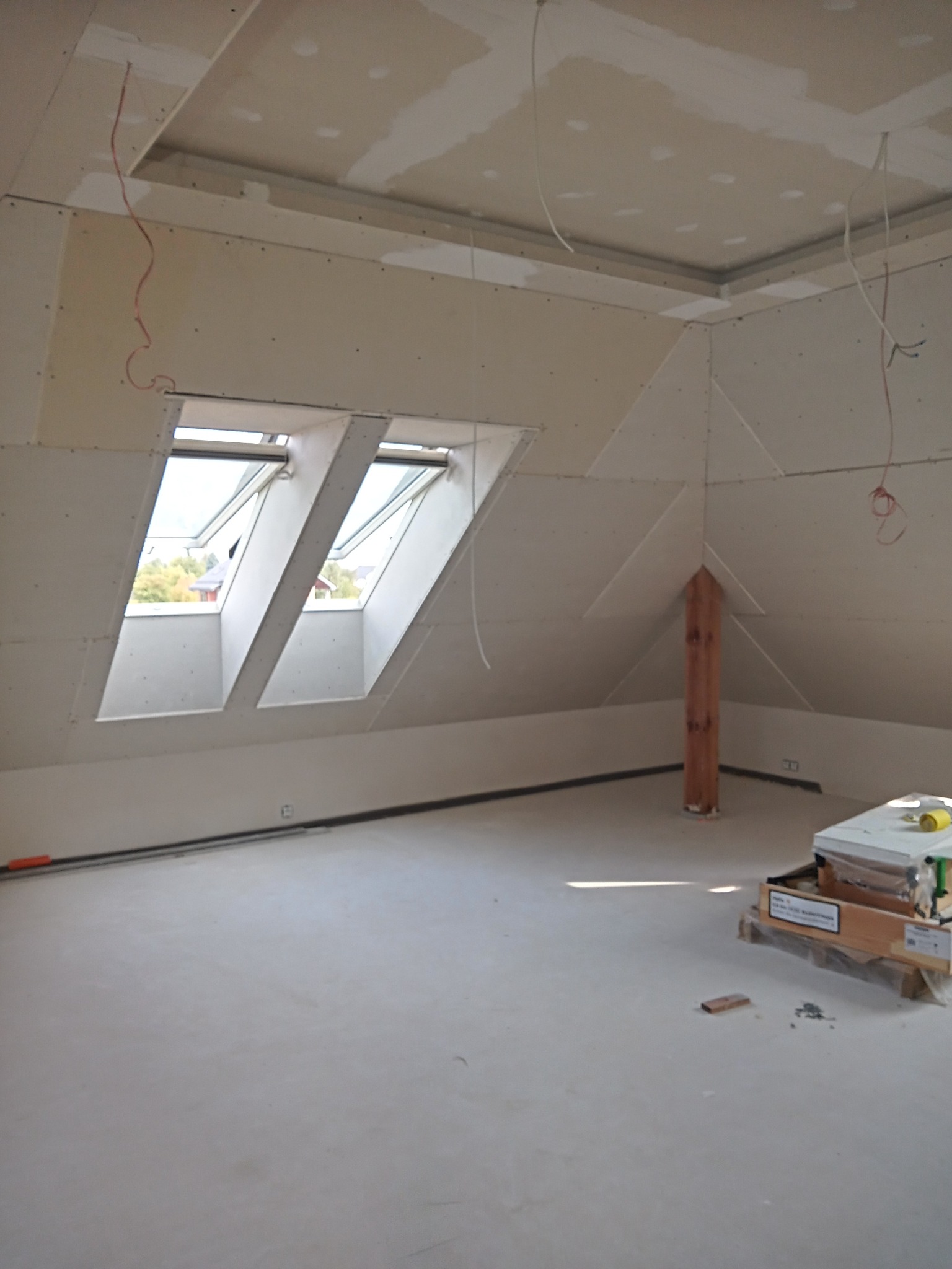 Poddasze w trakcie prac wykończeniowych: widoczne okna dachowe, zamontowane płyty gipsowo-kartonowe na skosach, drewniany słup podtrzymujący konstrukcję dachu oraz zwisające przewody elektryczne.