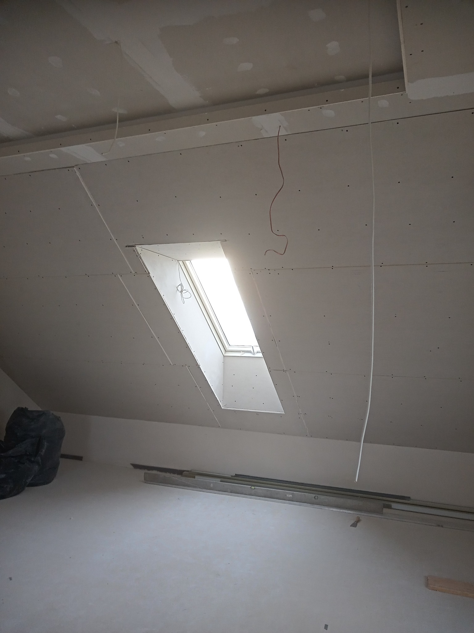 Poddasze w trakcie prac wykończeniowych: zamontowane płyty gipsowo-kartonowe na skosach i suficie, widoczne okno dachowe z wpadającym światłem, wiszące przewody elektryczne i częściowo zakryty...