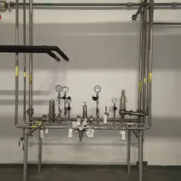 Instalacja przemysłowa z rur ze stali nierdzewnej, wyposażona w manometry i zawory, umieszczona w sterylnym, przemysłowym pomieszczeniu z szarym betonem na podłodze.