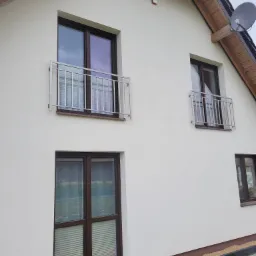 Elewacja domu z dwoma balkonami zabezpieczonymi metalowymi barierkami o minimalistycznym designie, widoczne okna z brązowymi ramami i satelita na dachu.