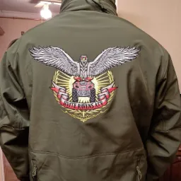 Plecy osoby w oliwkowej kurtce z dużym, kolorowym haftem przedstawiającym orła nad pojazdem ATV otoczonym wieńcem i napisem 'HVITE RAVENS ATV'.