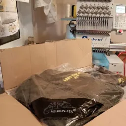 Nowa kurtka Helikon-Tex w plastikowym opakowaniu w kartonie, z maszyną do haftowania w tle.
