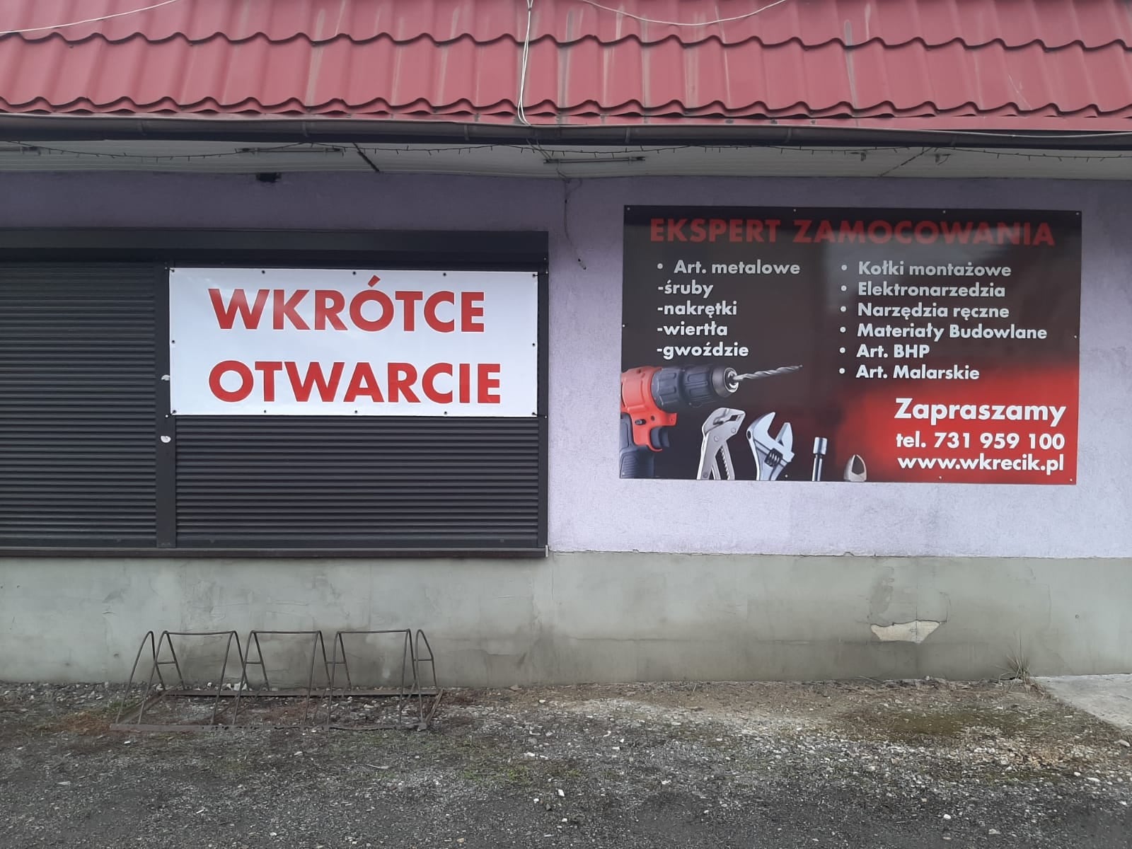 Fasada budynku z szyldem 'Wkrótce otwarcie' i reklamą 'Ekspert Zamocowania' oferującego artykuły metalowe, śruby, elektronarzędzia i materiały budowlane, widoczny stojak na rowery.