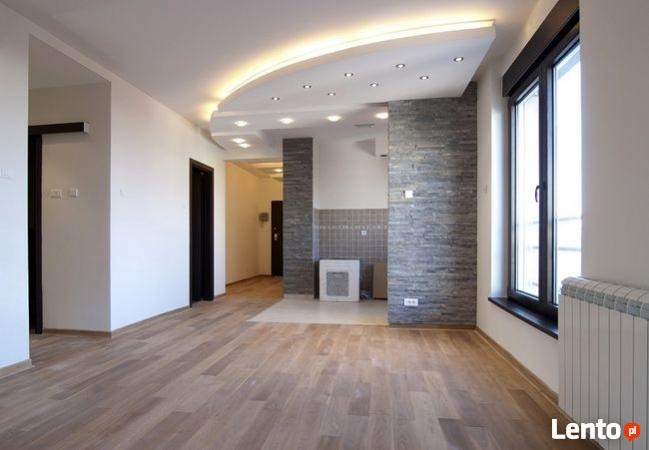 Nowoczesne wnętrze mieszkania: parkiet w odcieniach brązu, ściana z szarego kamienia dekoracyjnego, podwieszany sufit z oświetleniem LED, czarne ramy okienne, biały grzejnik.