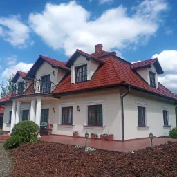 Elegancki, biały dom z czerwoną dachówką i balkonem z kutą balustradą, otoczony zadbanym ogrodem z korą i kwiatami, na tle błękitnego nieba z obłokami.