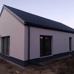 Nowoczesny dom jednorodzinny z jasną elewacją, ciemną podmurówką i grafitowym dachem, widoczny klimatyzator na ścianie, rynna odprowadzająca wodę