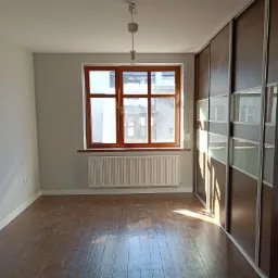 Pusty pokój po remoncie: odnowiona podłoga z ciemnego drewna, szare ściany, drewniane okno z grzejnikiem pod spodem, duża szafa przesuwna z ciemnego drewna i szkła.