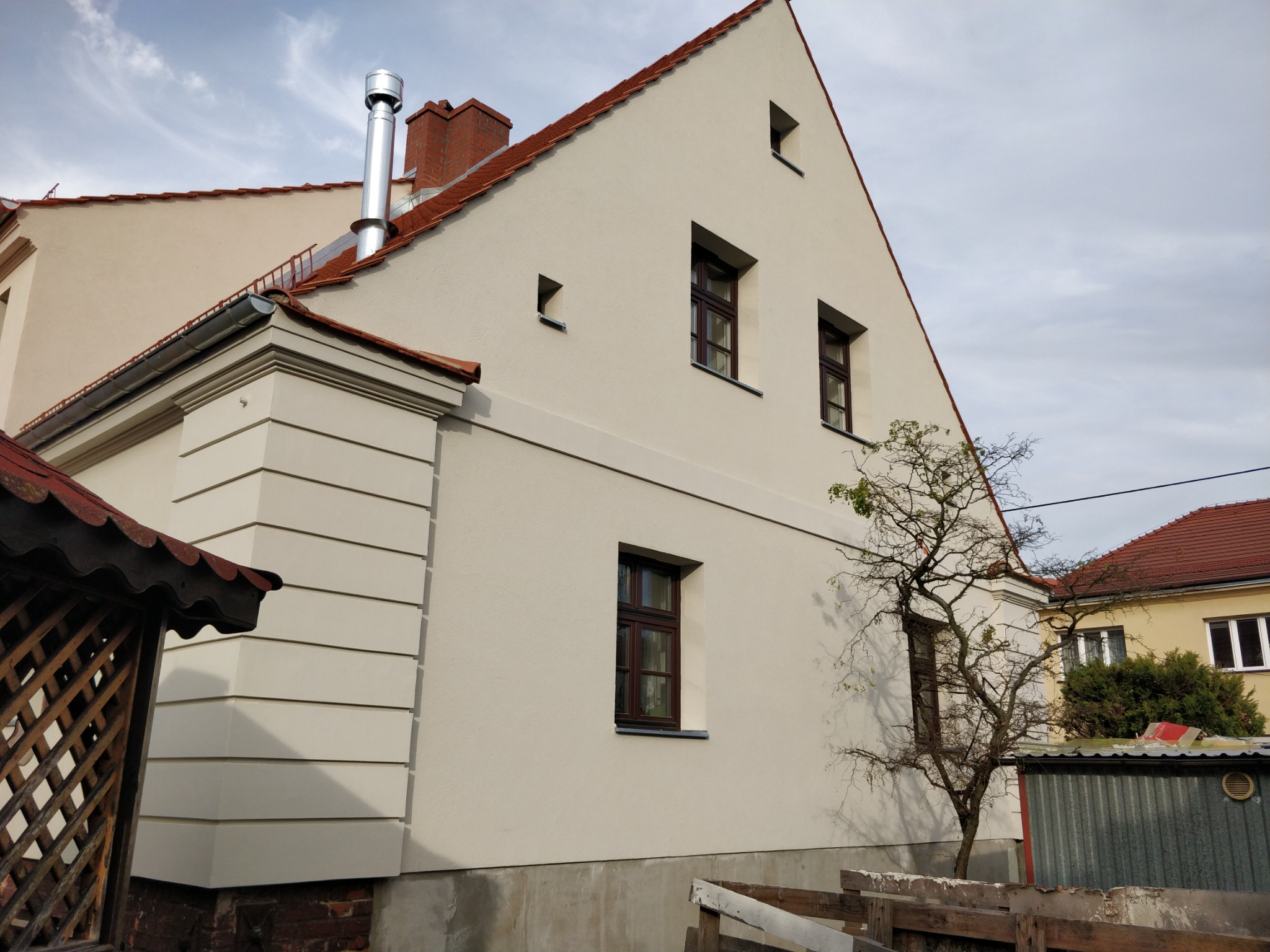Odnowiona elewacja budynku z kremową fasadą, brązowymi ramami okiennymi i czerwoną dachówką; widoczny komin ze stali nierdzewnej i niewielkie drzewo bez liści.
