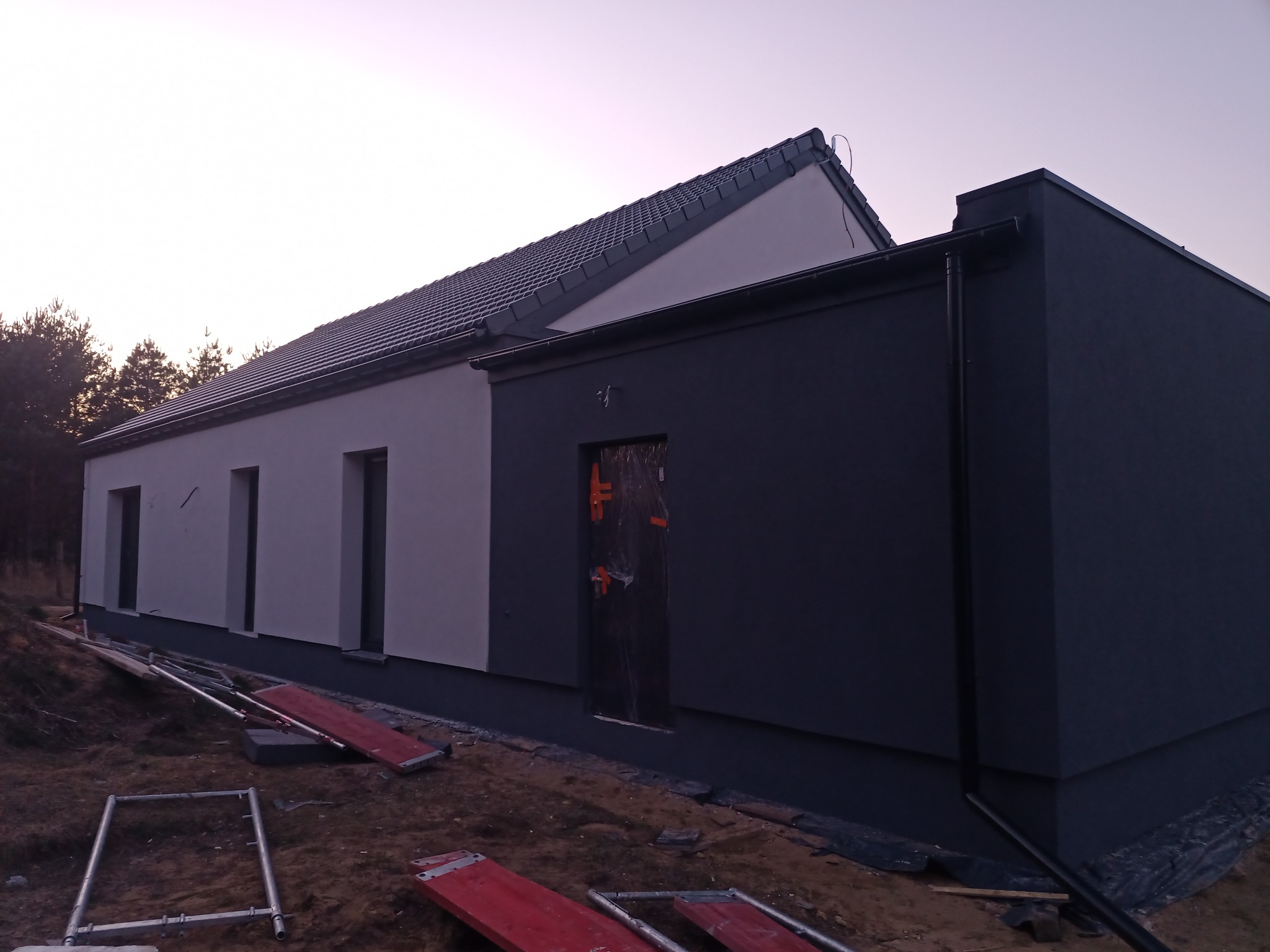 Nowoczesny dom z dwukolorową elewacją: jasnoszara i grafitowa, z widocznymi rusztowaniami i zabezpieczonym wejściem folią.