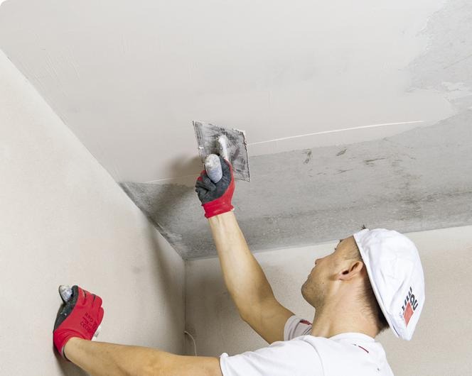 Mężczyzna w czerwonych rękawicach i białej czapce wygładza szpachlą sufit w rogu pomieszczenia, widoczna surowa powierzchnia ściany i fragment sufitu przed obróbką.