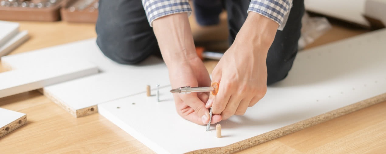 Montaż białej płyty meblowej za pomocą śrubokręta i drewnianych kołków na podłodze z paneli.