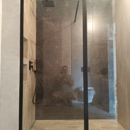 VP Remont Volodymyr Yavorskyi - Nowoczesna kabina prysznicowa z czarnymi profilami i deszczownicą, widoczne płytki w odcieniach szarości i beżu, odbicie fotografa w szkle.