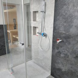 VP Remont Volodymyr Yavorskyi - Nowoczesna kabina prysznicowa z odpływem liniowym, szare płytki imitujące kamień, widoczna instalacja prysznicowa z niebieskim zabezpieczeniem na baterii, w tle fragment sauny w trakcie budowy.