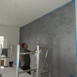 VP Remont Volodymyr Yavorskyi - Pracownik nanosi dekoracyjną, szarą strukturę na ścianę, zabezpieczoną niebieską taśmą malarską, używając szpachelki; widoczne drabiny, stół roboczy z narzędziami i puszkami farby.