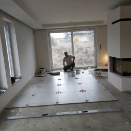 VP Remont Volodymyr Yavorskyi - Układanie jasnych, kwadratowych płytek podłogowych z użyciem poziomicy i czerwonych krzyżyków dystansowych w przestronnym, jasnym salonie z kominkiem; glazurnik w trakcie pracy.