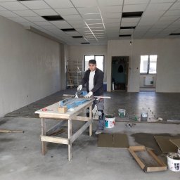 VP Remont Volodymyr Yavorskyi - Mężczyzna w roboczym ubraniu tnie płytki ceramiczne na drewnianym stole w przestronnym, surowym wnętrzu z widocznymi elementami przygotowania podłoża i pustą przestrzenią pod sufitem kasetonowym.