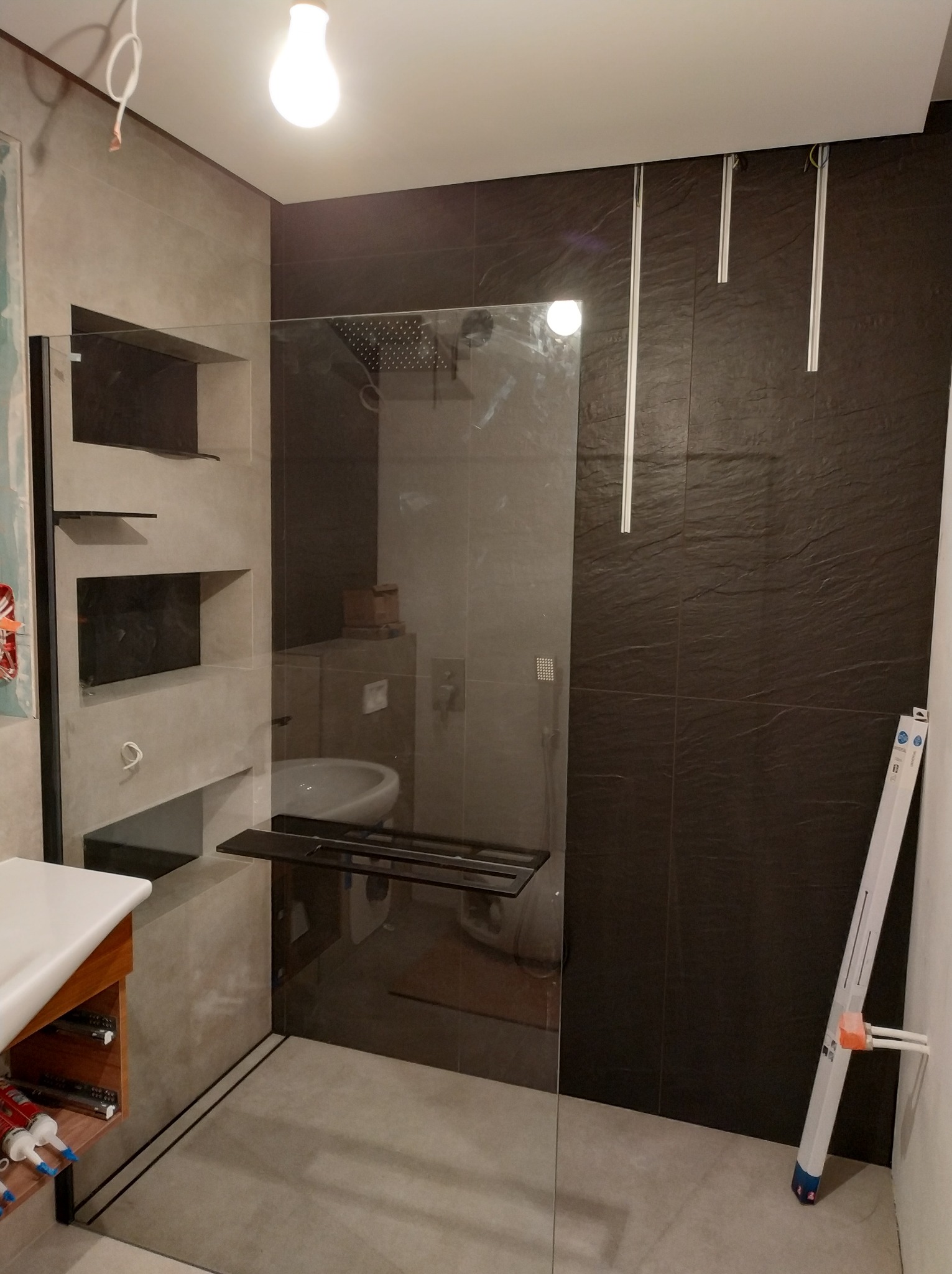 Wnętrze łazienki w trakcie remontu z widoczną kabiną prysznicową ze szklaną ścianą, półkami w ścianie, ciemnymi płytkami na jednej ze ścian oraz materiałami budowlanymi opartymi o ścianę.