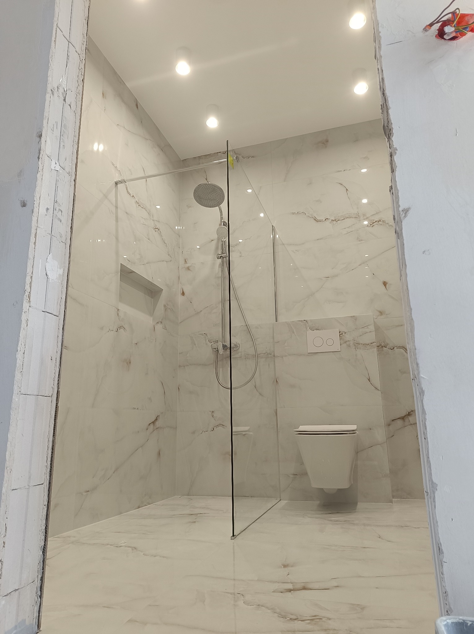 Wykończona łazienka z kabiną prysznicową typu walk-in, wiszącą toaletą i marmurowymi płytkami na ścianach i podłodze, widok z zewnątrz przez surowy otwór drzwiowy.