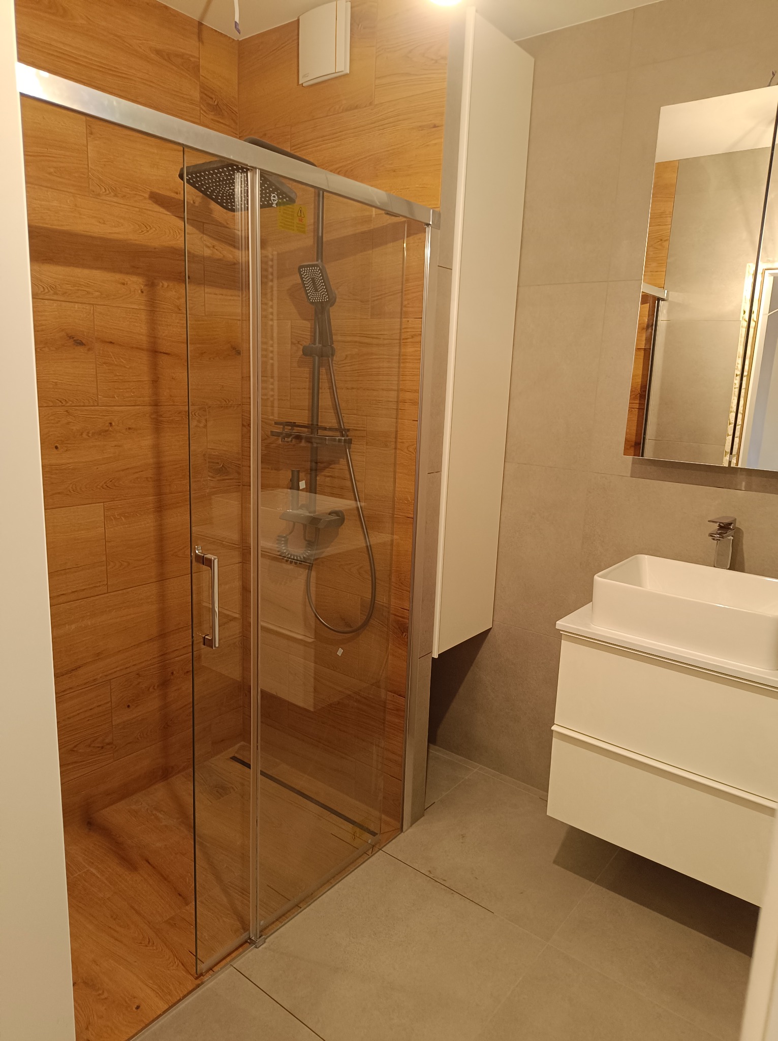 Nowoczesna łazienka z kabiną prysznicową typu walk-in, wykończona płytkami imitującymi drewno i szarymi płytkami, biała szafka z umywalką, lustro prostokątne.