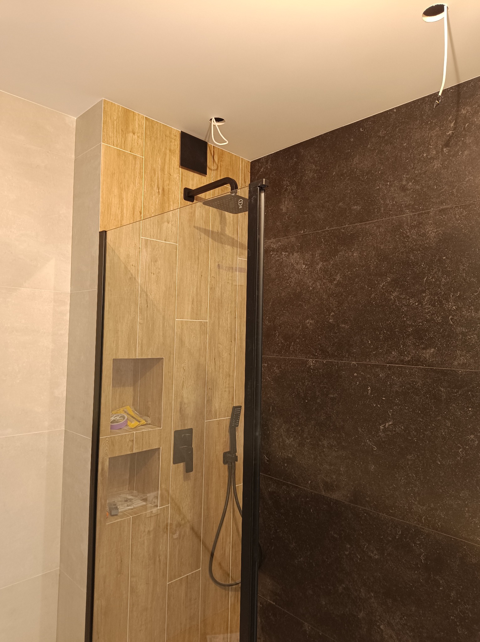 Nowoczesna kabina prysznicowa z brązowymi płytkami imitującymi drewno, wnęki na kosmetyki, czarna armatura i częściowo widoczne, ciemne płytki na ścianie po prawej stronie. Widoczne niedokończone...
