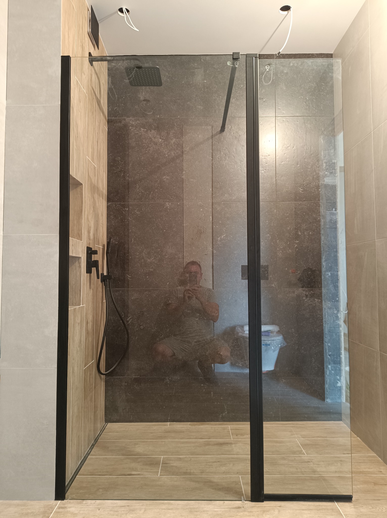 Nowoczesna kabina prysznicowa z czarnymi profilami i deszczownicą, widoczne płytki w odcieniach szarości i beżu, odbicie fotografa w szkle.