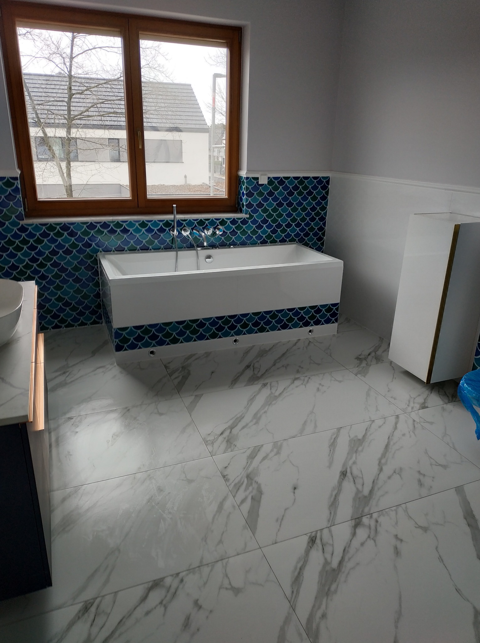 Nowoczesna łazienka z wanną obudowaną płytkami w kształcie rybiej łuski, marmurową podłogą i drewnianym oknem z widokiem na dom.