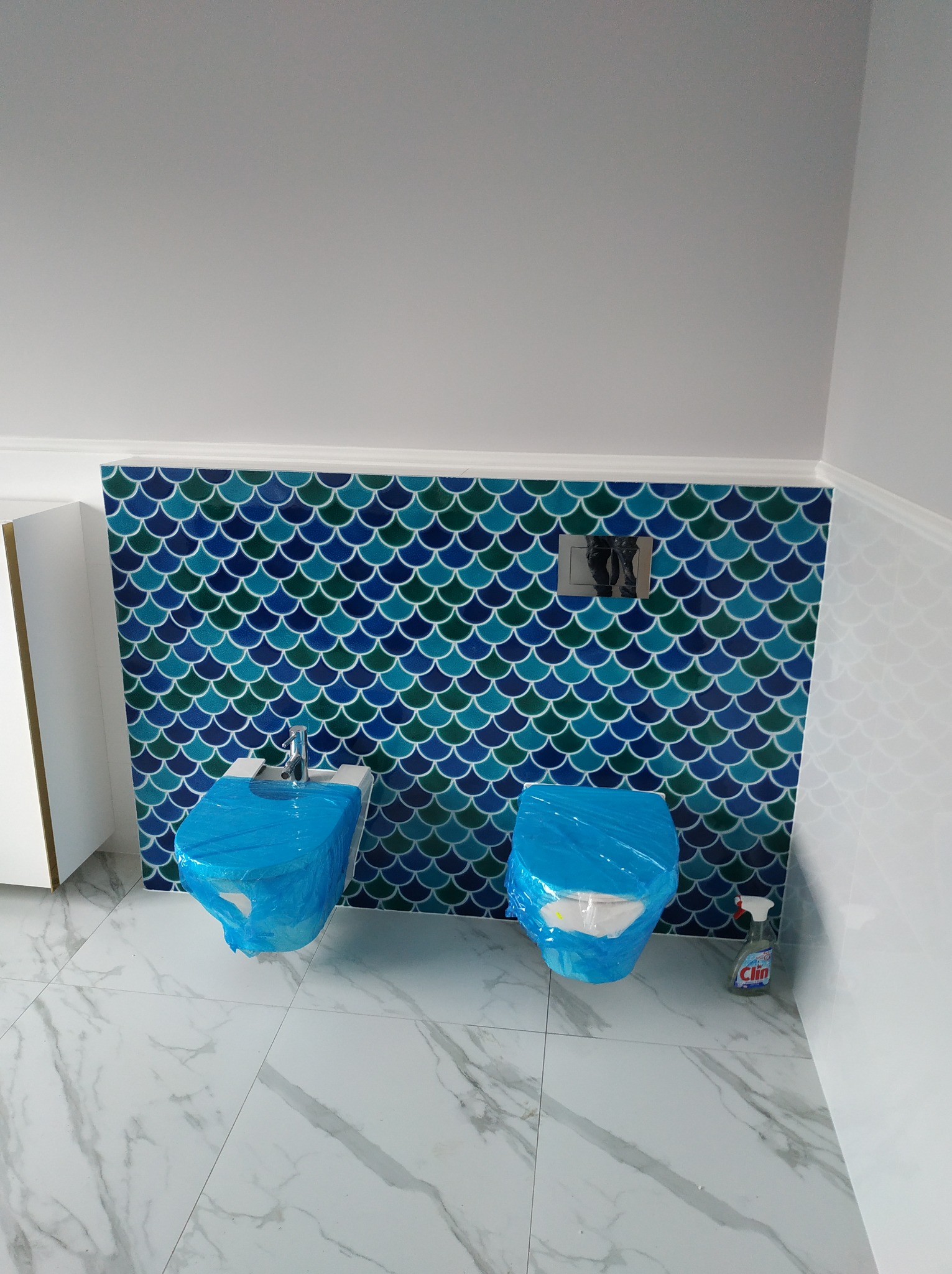 Bidet i toaleta w trakcie zabezpieczania folią ochronną, przed położeniem płytek o wzorze rybiej łuski w odcieniach niebieskiego i zielonego, na podłodze płytki imitujące marmur, widoczny środek...